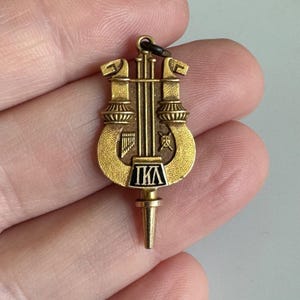 May include: A gold-colored pendant featuring a lyre design with the Greek letters ΠΚΑ in a black and white rectangular box. The pendant has a small loop at the top for attaching to a chain or necklace.