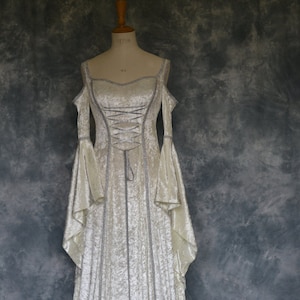Puede incluir: Un vestido de terciopelo blanco con ribetes de encaje plateado y un corpi&ntilde;o con cordones. El vestido tiene una falda larga y fluida y mangas acampanadas.