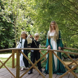 Puede incluir: Una familia vestida con trajes medievales se encuentra en un puente de madera en un entorno boscoso. La mujer lleva un vestido largo verde y blanco con detalles dorados. El niño lleva un traje negro y verde con una espada. La niña lleva un vestido verde y blanco con una corona de flores.