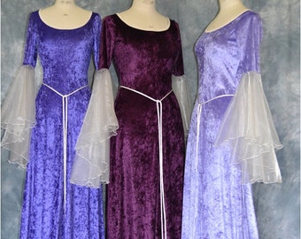 May include: Three long, flowing velvet dresses in shades of purple, lavender, and blue. Each dress has a white sash and sheer, white, ruffled sleeves. The dresses are displayed on mannequins against a gray background. The website address "www.frockfollies.com" is printed at the bottom of the image.