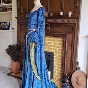 K&ouml;nnte beinhalten: Ein langes, blaues Samtkleid mit einem goldenen G&uuml;rtel und gr&uuml;nem Samtbesatz. Das Kleid befindet sich auf einer Schaufensterpuppe und wird vor einem Kamin ausgestellt.