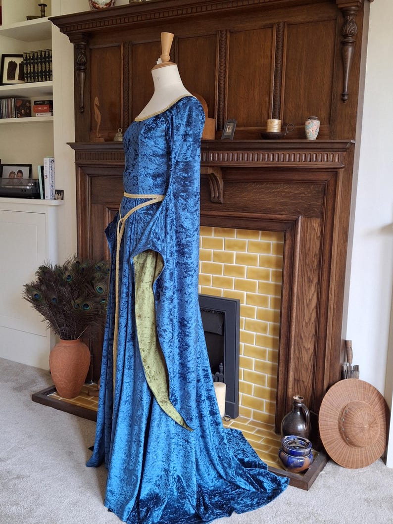 K&ouml;nnte beinhalten: Ein langes, blaues Samtkleid mit einem goldenen G&uuml;rtel und gr&uuml;nem Samtbesatz. Das Kleid befindet sich auf einer Schaufensterpuppe und wird vor einem Kamin ausgestellt.