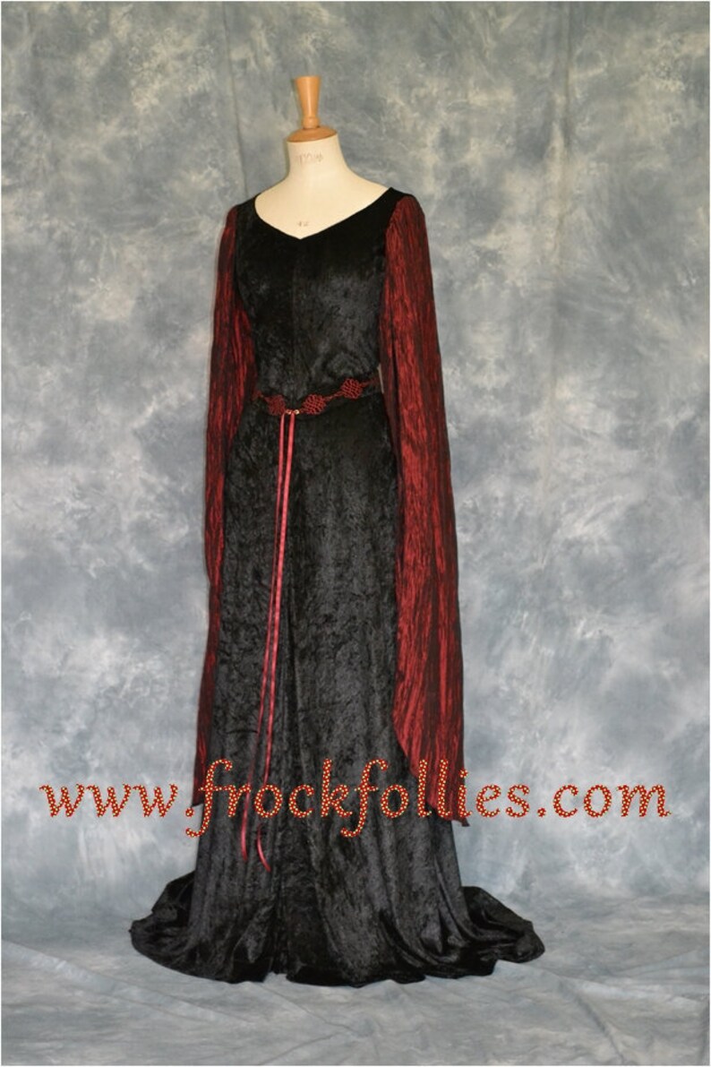 Op de afbeelding: Een zwarte fluwelen jurk met lange mouwen en een rode fluwelen rand. De jurk heeft een rode lintgordel met een decoratieve gesp. De jurk staat op een mannequin tegen een grijze achtergrond. www.frockfollies.com