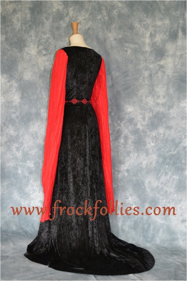 Op de afbeelding: Een zwarte fluwelen jurk met lange rode mouwen en een zwarte riem met een decoratieve gesp. De jurk heeft een sleep en wordt getoond op een mannequin. De tekst "www.frockfollies.com" is zichtbaar onderaan de afbeelding.