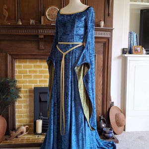 K&ouml;nnte beinhalten: Ein langes, blaues Samtkleid mit einem goldenen G&uuml;rtel. Das Kleid hat lange &Auml;rmel und einen runden Ausschnitt. Das Kleid befindet sich auf einer Schaufensterpuppe vor einem Kamin.