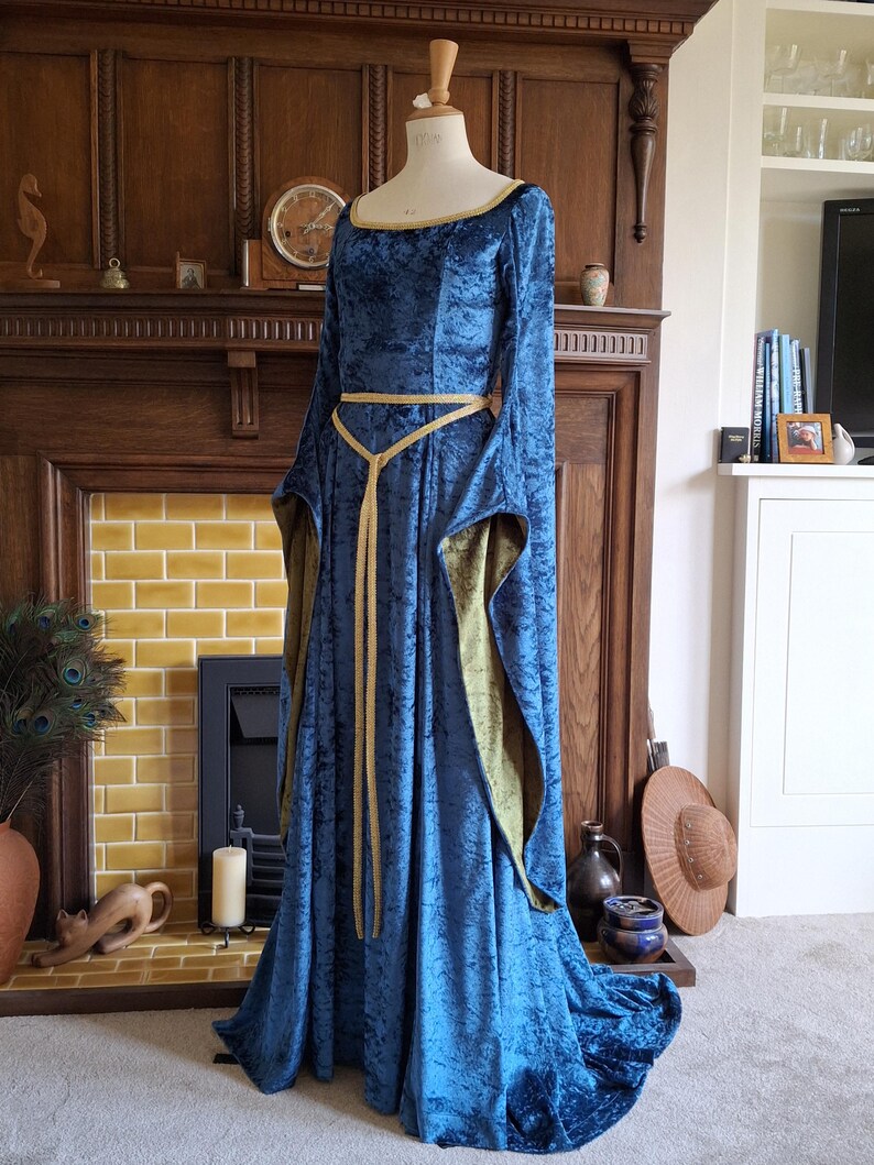K&ouml;nnte beinhalten: Ein langes, blaues Samtkleid mit einem goldenen G&uuml;rtel. Das Kleid hat lange &Auml;rmel und einen runden Ausschnitt. Das Kleid befindet sich auf einer Schaufensterpuppe vor einem Kamin.