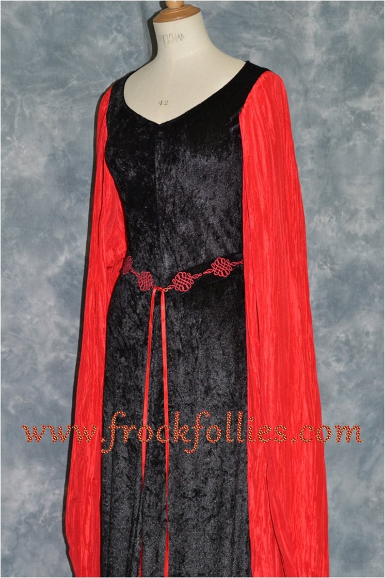 Op de afbeelding: Een zwarte fluwelen jurk met lange rode mouwen en een rode lintgordel. De gordel heeft een decoratief ontwerp van rode knopen. De jurk is op een mannequin en de tekst "www.frockfollies.com" is zichtbaar onderaan de afbeelding.