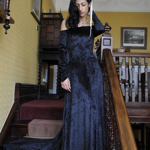 May include: A woman in a long, dark blue velvet dress with a white ribbon tied around her neck stands on a staircase. The dress has a ruffled neckline and long sleeves. The woman is looking down at the floor.