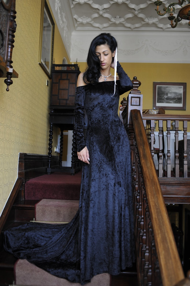 May include: A woman in a long, dark blue velvet dress with a white ribbon tied around her neck stands on a staircase. The dress has a ruffled neckline and long sleeves. The woman is looking down at the floor.