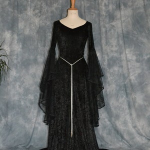 May include: A black velvet gown with long sleeves and a lace overlay. The gown has a fitted bodice and a flowing skirt. A silver cord belt cinches the waist.