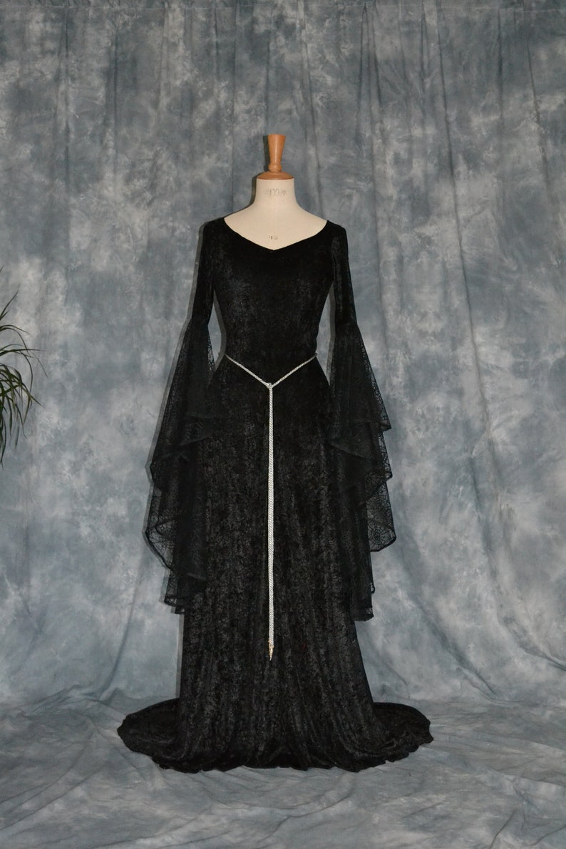 May include: A black velvet gown with long sleeves and a lace overlay. The gown has a fitted bodice and a flowing skirt. A silver cord belt cinches the waist.