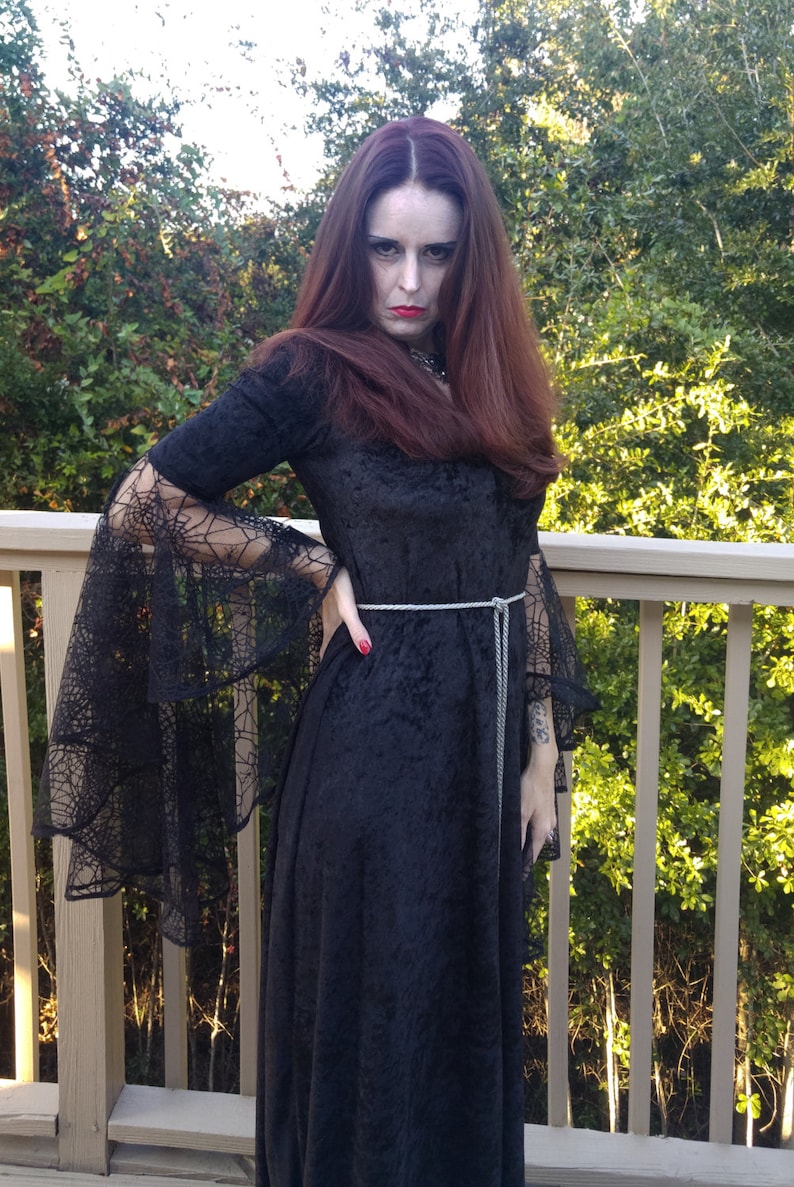 May include: A woman in a black velvet dress with spiderweb lace sleeves and a silver belt. She is standing in front of a wooden railing with green foliage behind her.