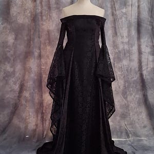Puede incluir: Un vestido negro largo hasta el suelo con escote de hombros descubiertos y mangas largas y anchas de encaje. El corpiño y la falda están confeccionados con una tela negra sólida con un sutil patrón floral tonal en toda la prenda.