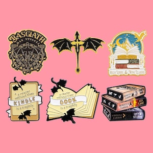 Könnte beinhalten: Eine Sammlung von Emaille-Anstecknadeln mit Fantasy-Motiven. Designs umfassen einen Drachen, ein Schild, einen Bücherstapel und Anstecknadeln mit dem Text "a reader without their KINDLE is a tragedy" und "a reader without their BOOK is a tragedy".