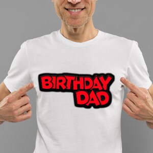 K&ouml;nnte beinhalten: Ein wei&szlig;es T-Shirt mit dem Aufdruck "Birthday Dad" in roten und schwarzen Buchstaben.