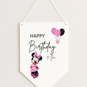 May include: White pennant-shaped wall hanging with a Minnie Mouse illustration and the text "Happy Birthday To You." Minnie Mouse is wearing a pink polka-dot dress and bow. Pink and black balloons are also featured.