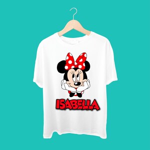 K&ouml;nnte beinhalten: Wei&szlig;es T-Shirt mit einer roten Schleife mit wei&szlig;en Punkten, einem Cartoon-Bild von Minnie Mouse und dem Namen "Isabella" in Rot.