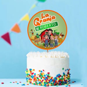 May include: A white cake with colourful sprinkles at the base, topped with a round cake topper. The topper features cartoon characters and the text "La Granja DE ROBERTO" in orange and green.