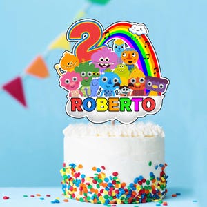 Könnte beinhalten: Ein weißer Kuchen mit bunten Streuseln am Boden. Ein Tortenaufsatz zeigt einen Regenbogen, Zeichentrickfiguren, die Zahl 2 und den Namen "ROBERTO" in bunten Buchstaben. Der Hintergrund ist hellblau.
