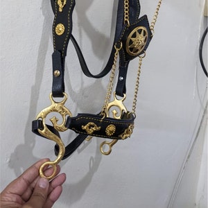 Op de afbeelding: Zwart lederen paardenhoofdstel met goudkleurige hardware en een decoratieve ketting. Het hoofdstel heeft een decoratief ontwerp met een kroon en de tekst "Horse Tack's".
