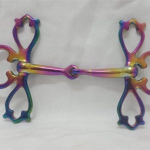 Op de afbeelding: Een regenboogkleurig paardenbit met een centraal gewricht en gebogen zijstukken. Het bit heeft een metallic afwerking en is ontworpen voor paardensport. Het logo "Horse Tack's" is linksboven zichtbaar.