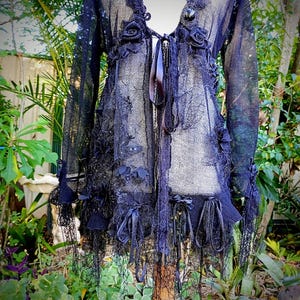 May include: A black, open-front lace jacket with long sleeves, adorned with fabric roses, bows, and dangling lace trim. The jacket features a tie closure and is displayed on a mannequin in a garden setting. The garment is a unique piece of apparel.