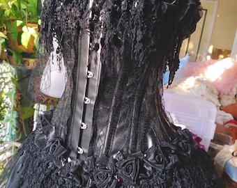 May include: A black corset with lace and satin rose embellishments. The corset has a busk closure in the front and is worn over a black tulle skirt.
