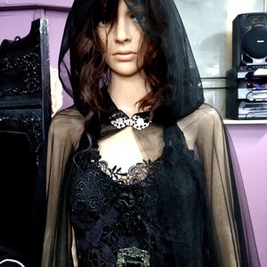 May include: A black sheer cape with a lace bodice and a large black ornate buckle. The cape has a hood and is made of a soft, flowing fabric.