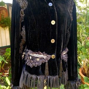 May include: A black velvet jacket with lace trim and gold buttons. The jacket has a ruffled hem and a belt with a beaded embellishment.
