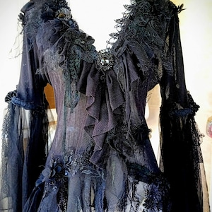 May include: Black gothic-style top with long sleeves, intricate lace, and ruffled detailing. The dark garment features a deep V-neck, embellished with decorative elements and a layered, textured design, creating a dramatic and elegant aesthetic.