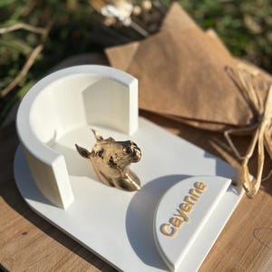 Può includere: Un elemento decorativo bianco a forma di cuore con una scultura di testa di cavallo dorata e il nome "Cayenne" in lettere dorate. Il pezzo ha un arco semicircolare ed è posto su una superficie di legno.