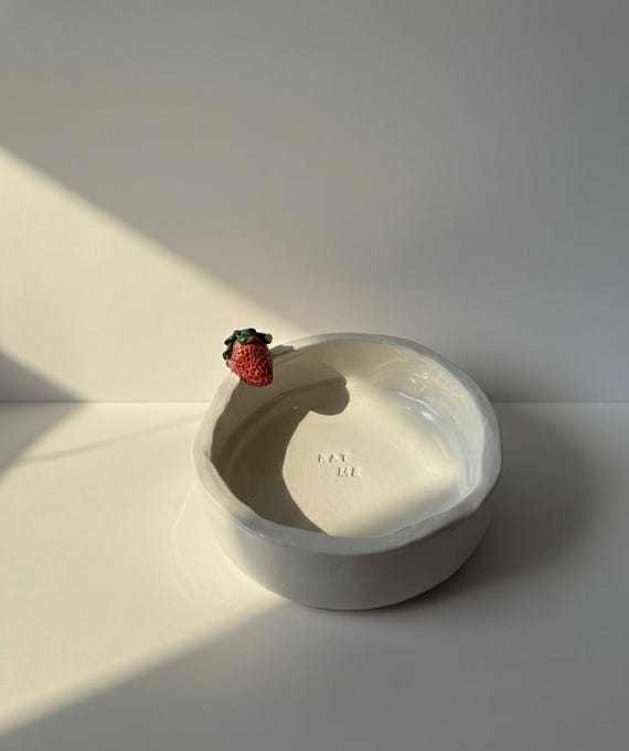 A sculptural ceramic strawberry bowl stamped 'EAT ME' — whimsical, artisan-made pottery with genuine visual wit that stops a scroll dead in its tracks.