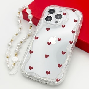 Puede incluir: Funda transparente para iPhone con un patrón de corazones rojos. La funda tiene un borde ondulado y una correa para teléfono de perlas y oro. La funda está junto a una caja roja.
