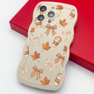 Peut inclure: Une coque de téléphone de couleur crème aux bords ondulés présentant un motif répétitif de feuilles d'érable orange, de nœuds à carreaux marron et blancs, de nœuds couleur crème et de gobelets à café jetables marron et blancs.