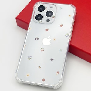 May include: Clear phone case with a floral and fruit pattern, including strawberries, cherries, and small flowers. The case is on a red surface. The phone case has a clear bumper.