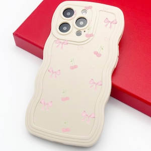 Op de afbeelding: Een beige telefoonhoesje met een golvende rand, versierd met roze strikken, kersen en hartjes. De hoes heeft een camera-uitsparing voor meerdere lenzen. De hoes ligt op een rode ondergrond.