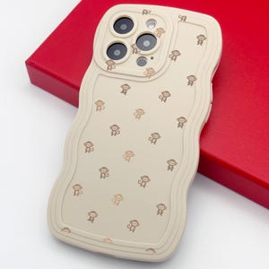 May include: Beige phone case with a wavy border and a pattern of small brown monkey illustrations. The case has cutouts for the phone's camera and buttons. The case is on a red surface.