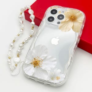 Könnte beinhalten: Transparente Handyhülle mit weißem Blumendesign und Perlenkette. Die Hülle hat ein klares Design mit weißen Blumen und goldenen Akzenten. Die Perlenkette besteht aus weißen Perlen und goldenen Perlen.