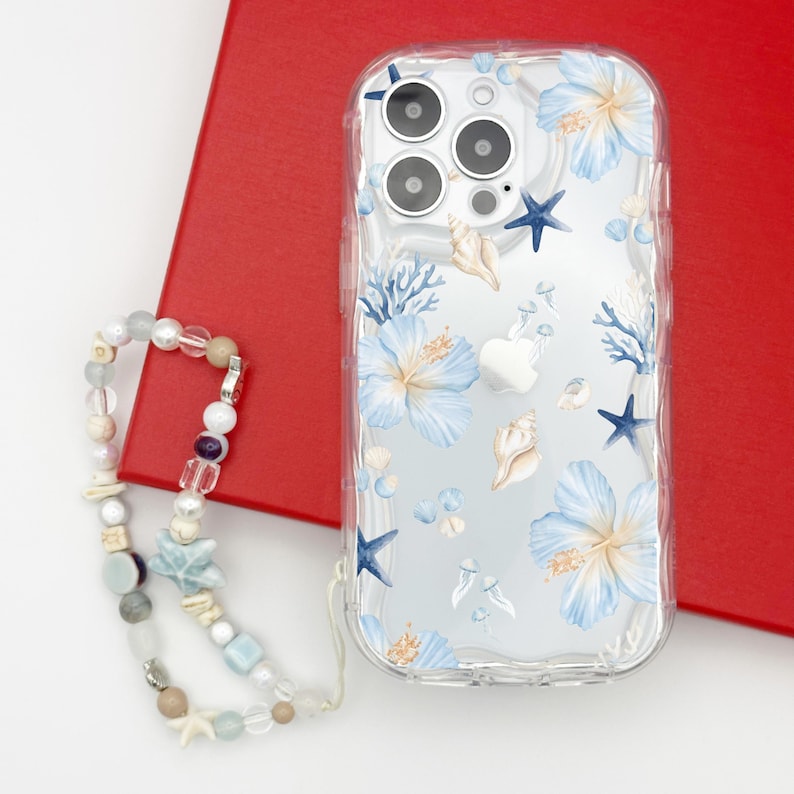K&ouml;nnte beinhalten: Transparente Handyh&uuml;lle mit Blumen- und Muschelmuster. Die H&uuml;lle zeigt blaue Hibiskusbl&uuml;ten, Seesterne und Muscheln. Eine Perlen-Handy-Schlaufe mit verschiedenen Perlen und Anh&auml;ngern befindet sich daneben.