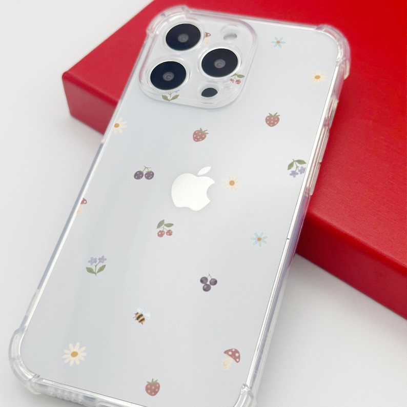 May include: Clear phone case with a whimsical design featuring strawberries, cherries, flowers, and a mushroom. The case has a transparent border and is decorated with small, colorful illustrations. The Apple logo is visible on the back.