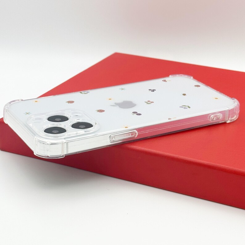 May include: Clear phone case with a decorative pattern of small, colorful shapes, including strawberries and cherries. The case has a clear bumper and covers the back of a phone with a triple-lens camera. The case is resting on a red surface.