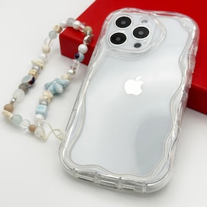 Peut inclure: Une coque de téléphone transparente avec un design ondulé et une bosse de caméra surélevée. La coque est sur une surface rouge et a une dragonne de téléphone perlée attachée.