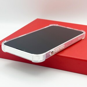 May include: A clear, protective phone case with a black screen, resting on a red rectangular box. The case has a transparent design with reinforced corners and button covers, offering a sleek and modern look. The phone screen is black.