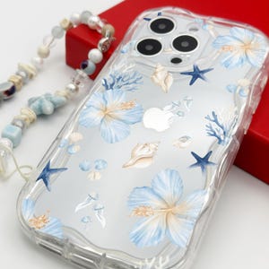 K&ouml;nnte beinhalten: Transparente Handyh&uuml;lle mit Strandmotiv. Sie zeigt blaue Hibiskusbl&uuml;ten, Muscheln, Seesterne und Korallen. Eine Perlen-Handschlaufe mit blauen Akzenten ist angebracht. Die H&uuml;lle ist f&uuml;r ein Smartphone konzipiert.