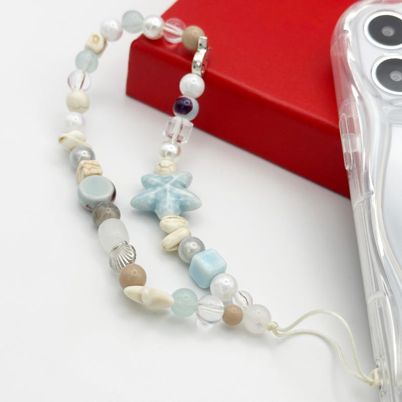 K&ouml;nnte beinhalten: Ein mit Perlen besetzter Handy-Anh&auml;nger mit einer Vielzahl von Perlen in Wei&szlig;, Beige, Blau und Transparent. Der Anh&auml;nger enth&auml;lt eine hellblaue, sternf&ouml;rmige Perle und einen silberfarbenen Verschluss. Der Anh&auml;nger ist an einer transparenten Handyh&uuml;lle befestigt.