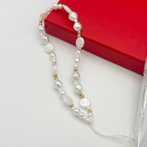 Op de afbeelding: Een telefoonbedel van witte parels en een gouden ketting met een wit touwtje.