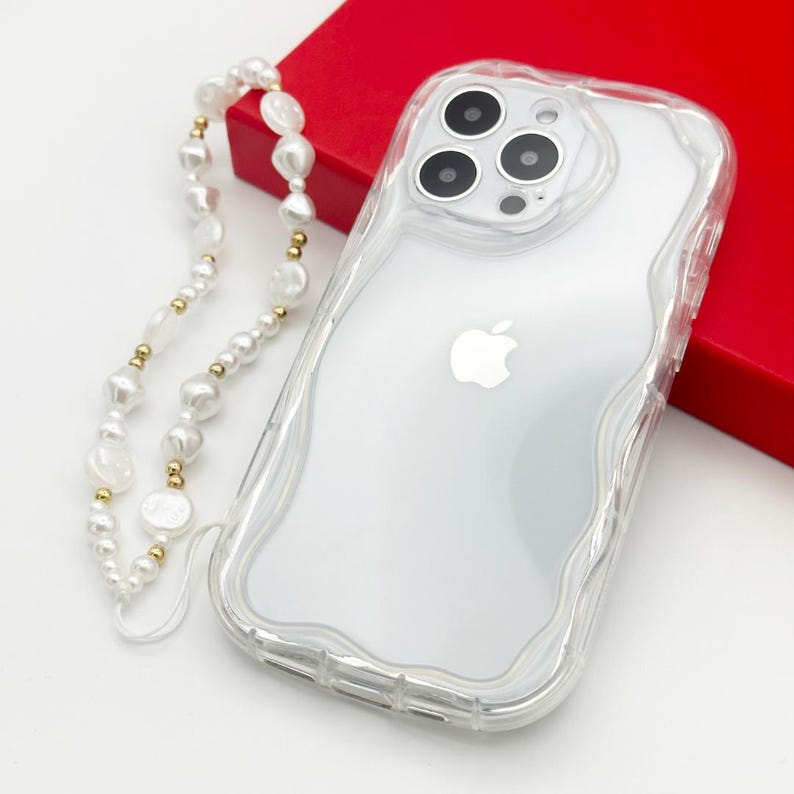 Op de afbeelding: Een transparante telefoonhoes met een golvend ontwerp en een verhoogde camerabult. De hoes wordt geleverd met een witte parelketting met gouden accenten.