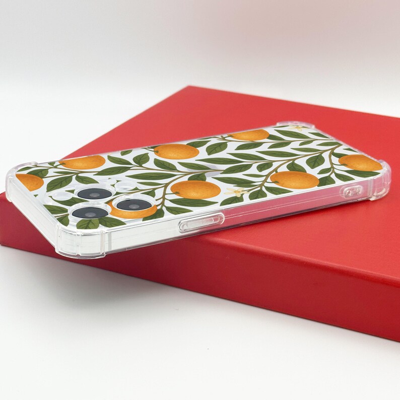 May include: A clear phone case featuring a pattern of orange fruit and green leaves on a white background. The case is rectangular and has a clear border. The phone case is resting on a red surface.