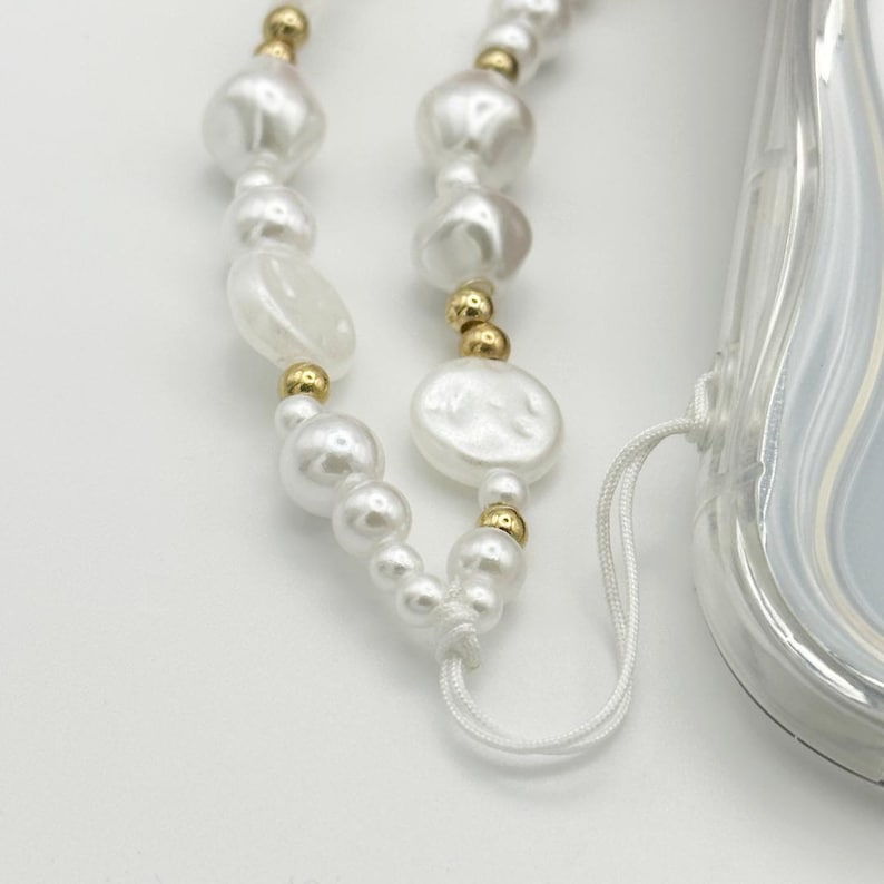 Op de afbeelding: Een witte telefoonhoes met een telefoonband van parels en gouden kralen. De band heeft een wit koord met een knoop aan het einde.
