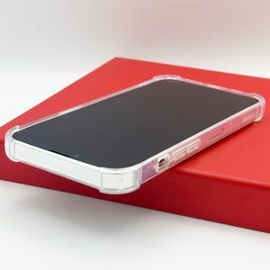 May include: A clear protective phone case with a black screen, resting on a red surface. The case has a transparent design with reinforced corners and side buttons. The phone case is designed to protect the phone from damage.